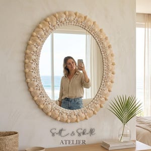 May include: A circular mirror, framed with seashells, is mounted on a white wall. The mirror reflects a person taking a photo. The text "Salt & Shell ATELIER" is below. A bedroom with a bed and window is visible in the background.