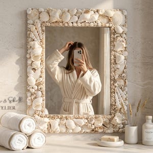 May include: A rectangular mirror with a frame intricately decorated with various seashells. The mirror reflects a person in a white robe. The scene includes rolled white towels, a soap dish, and a bottle labeled "Coastal Spa."