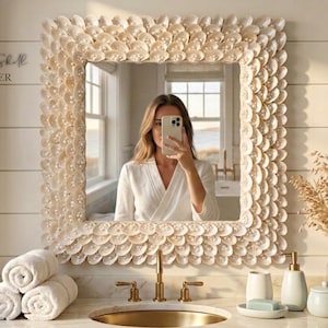 May include: A square mirror with a frame decorated with shell-shaped elements. The mirror is above a bathroom sink with gold fixtures and white towels. The text "Salt & Shell ATELIER" is visible in the upper left corner.