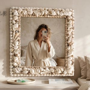 Puede incluir: Espejo cuadrado con un marco intrincadamente decorado con varias conchas marinas. El espejo refleja a una persona haciéndose un selfie. El texto "Salt & Shell ATELIER" está en la esquina superior izquierda. El espejo está sobre una cómoda blanca.
