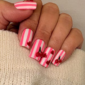 May include: A hand with painted fingernails. The nails are white with pink vertical stripes. Some nails have red heart and cherry designs. The thumb nail has pink and white stripes with red accents.