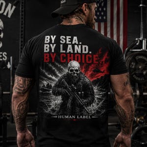 以下が含まれることがあります： 「BY SEA. BY LAND. BY CHOICE.」の文字と、ライフルを持った骸骨顔の兵士のグラフィックがプリントされた黒いTシャツ。「HUMAN LABEL」の文字も含まれています。