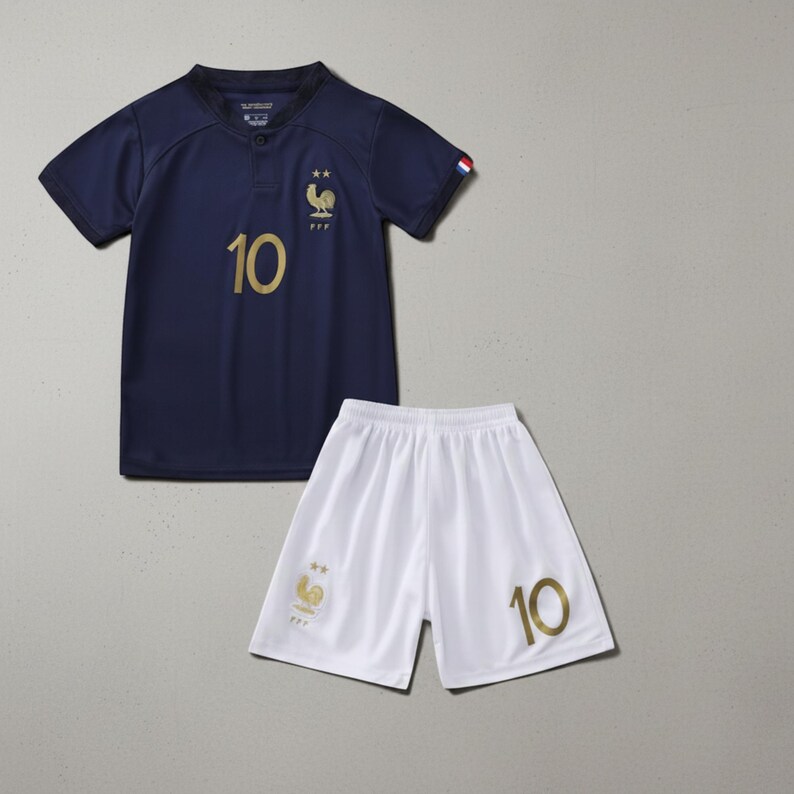 Pode incluir: Um conjunto de camisa e cal&ccedil;&atilde;o de futebol azul marinho. A camisa apresenta o n&uacute;mero 10 em dourado, um emblema de galo dourado e as letras FFF. O cal&ccedil;&atilde;o branco tamb&eacute;m exibe o n&uacute;mero 10 em dourado.
