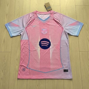 以下が含まれることがあります： ピンクとライトブルーのサッカーユニフォーム、Vネックカラー。前面にはSpotifyのロゴとチームの紋章があります。袖はピンクからライトブルーに変わるグラデーションデザインです。胸にはナイキのロゴが見えます。
