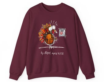 May include: A maroon crewneck sweatshirt featuring a colorful, scribbled illustration of a turkey looking at a calendar marked with the text NOV 25. Below the graphic, the text reads by Nigel, aged 6 1/2 in a white, hand-drawn style font.