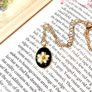 May include: Gold-toned necklace with an oval pendant featuring a white flower on a black background. The pendant hangs from a delicate chain. The necklace is displayed on a book with printed text.
