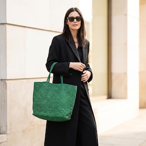 May include: A bright green woven tote bag with matching handles. The bag is held by a person wearing a black coat and sunglasses. The bag has a rectangular shape and a textured surface.
