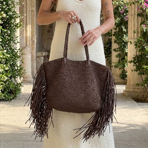 May include: A brown woven tote bag with long fringe detailing. The bag has braided handles and leather accents. The bag is held by a person wearing a white dress.