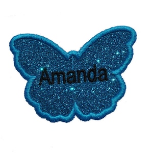 Pode incluir: Uma borboleta azul com glitter, contorno preto e o nome "Amanda" escrito no centro.