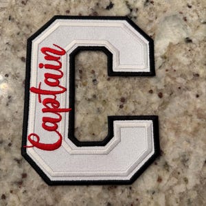 May include: White embroidered letter "C" patch with black and white trim. The word "Captain" is embroidered in red script on the left side of the letter. The patch is likely for a jacket or uniform.