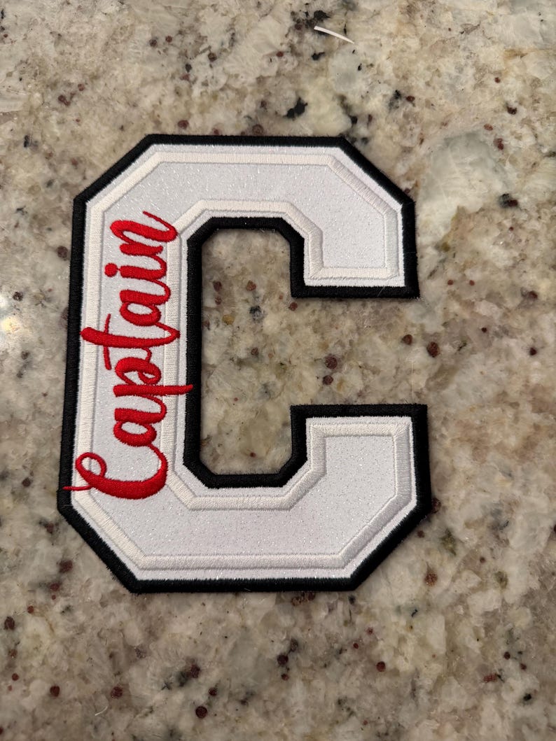 May include: White embroidered letter "C" patch with black and white trim. The word "Captain" is embroidered in red script on the left side of the letter. The patch is likely for a jacket or uniform.