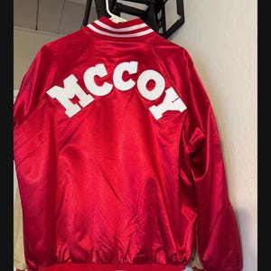 May include: A red satin jacket with the name "MCCOY" in white block letters across the back. The jacket has white stripes on the collar, cuffs, and waistband.
