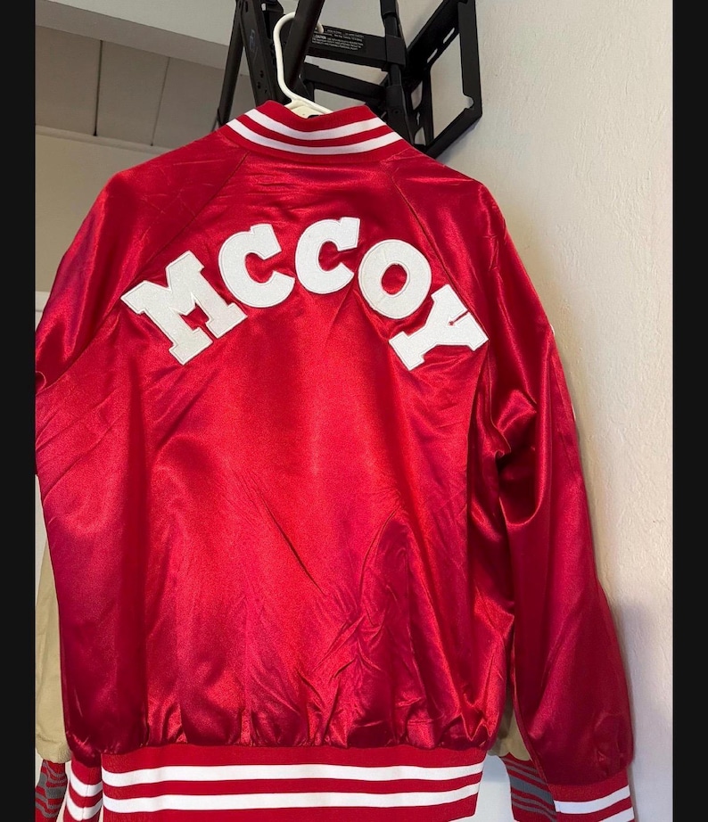 May include: A red satin jacket with the name "MCCOY" in white block letters across the back. The jacket has white stripes on the collar, cuffs, and waistband.
