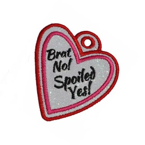 Peut inclure: Un patch en forme de cœur blanc avec une bordure rose et rouge. Le patch porte le texte "Brat No! Spoiled Yes!" en noir.