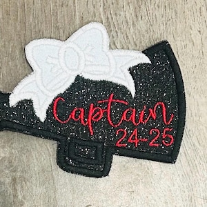 Peut inclure: Mégaphone noir pailleté avec un nœud blanc et une broderie rouge qui dit "Captain 24-25".