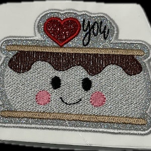 Pode incluir: Patch bordado com um s'more sorridente com bochechas rosadas, olhos pretos e uma camada de chocolate marrom. Um coração vermelho brilhante e a palavra "you" estão acima do s'more. O patch tem uma borda prateada brilhante.