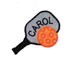 May include: A silver and black pickleball paddle with the name "CAROL" written on it. A bright orange pickleball is next to the paddle.