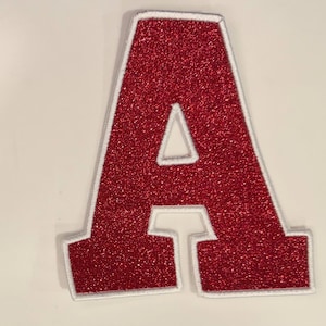 May include: A red glitter letter "A" patch with a white border. The patch is a capital letter "A" with a triangular cutout in the centre. The patch is likely for clothing or accessories.