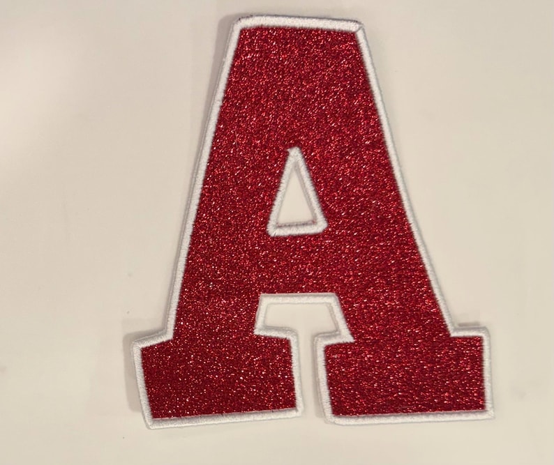 May include: A red glitter letter "A" patch with a white border. The patch is a capital letter "A" with a triangular cutout in the centre. The patch is likely for clothing or accessories.