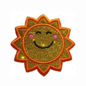 Pode incluir: Um sol de glitter dourado com contorno laranja e um rosto sorridente. O sol tem bochechas cor-de-rosa.