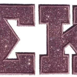Könnte beinhalten: Zwei gestickte griechische Buchstaben, Sigma und Kappa, mit einem dunkelmalvenfarbenen Glitzer-Finish. Jeder Buchstabe hat einen dicken, gestickten Rand aus passendem malvenfarbenem Garn, was für eine strukturierte Optik sorgt.