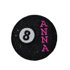 Puede incluir: Bola de billar 8 negra y blanca con purpurina rosa y el nombre "ANNA" bordado verticalmente en el lado derecho.