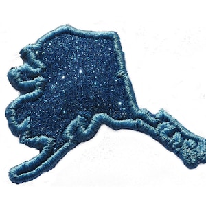 May include: A blue glitter outline of the state of Alaska. The outline is made of a sparkly blue fabric.