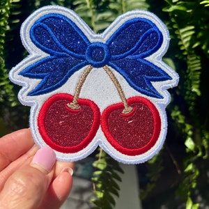 May include: Embroidered patch featuring a pair of red cherries with glitter accents, connected by brown stems. A blue glitter bow sits above the cherries, outlined in white. The patch has a white border.