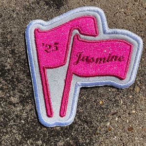May include: A pink glitter patch in the shape of two flags. The flags have a white border with blue stitching. One flag reads "'25" and the other reads "Jasmine" in a cursive font.