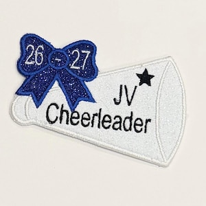 Op de afbeelding: Een witte glitterende patch in de vorm van een megafoon met een blauwe glitterende strik aan de bovenkant. De strik toont de cijfers 26-27. De megafoon bevat de zwarte tekst JV en Cheerleader met een klein zwart sterretje boven de letters JV.