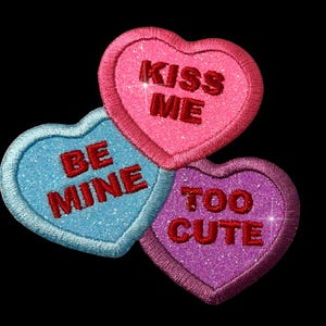 Pode incluir: Três corações de conversa de doces em forma de coração com glitter. O coração rosa diz "Kiss Me", o coração azul diz "Be Mine" e o coração roxo diz "Too Cute".