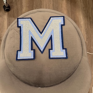 May include: A large, embroidered letter "M" patch. The letter is silver with a light blue outline and a navy blue border. The patch is on a gray, rounded surface.