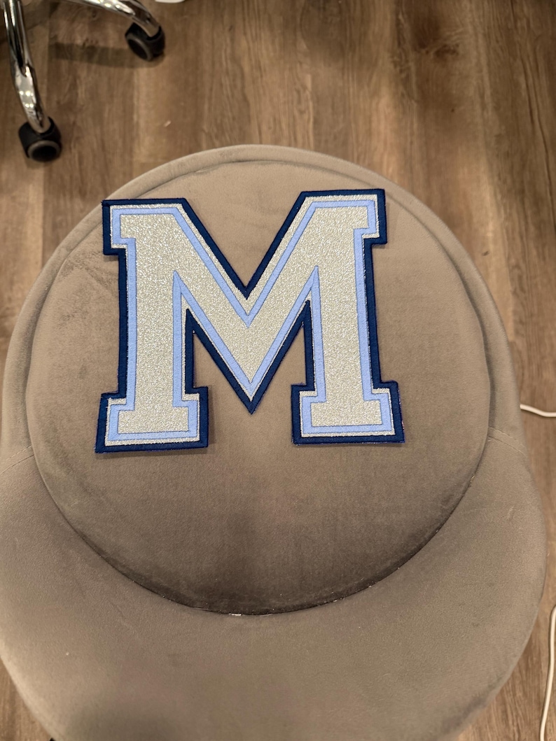 May include: A large, embroidered letter "M" patch. The letter is silver with a light blue outline and a navy blue border. The patch is on a gray, rounded surface.