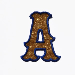 Könnte beinhalten: Ein goldener Glitzerbuchstabe A mit blauem Umriss. Der Buchstabe ist in einer Schriftart im Vintage-Stil.