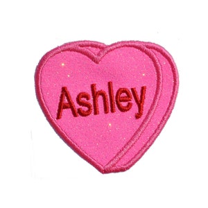 Pode incluir: Patch rosa em forma de coração com o nome "Ashley" bordado em vermelho.