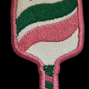 May include: Embroidered patch in the shape of a paddle, with a pink outline and handle. The paddle design features white, green, and pink stripes and a small pink heart. The patch has a glittery texture.