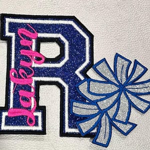 May include: A blue and white letter "R" patch with the name "Ruby" in pink script, and two silver and blue pom-poms. The patches have a glittery texture and are on a white background.