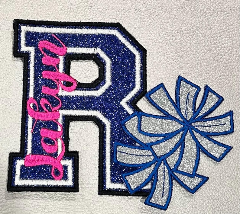May include: A blue and white letter "R" patch with the name "Ruby" in pink script, and two silver and blue pom-poms. The patches have a glittery texture and are on a white background.