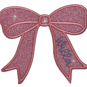May include: Pink glitter iron-on patch with a large bow and the name "Bella" written in blue thread.