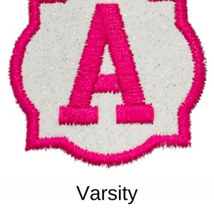 May include: A white felt patch with a pink felt outline in the shape of a rounded rectangle. The letter "A" is embroidered in pink felt in the center of the patch. The text "Varsity" is printed below the patch.