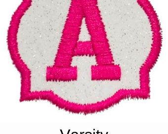 May include: A white felt patch with a pink felt outline in the shape of a rounded rectangle. The letter "A" is embroidered in pink felt in the center of the patch. The text "Varsity" is printed below the patch.