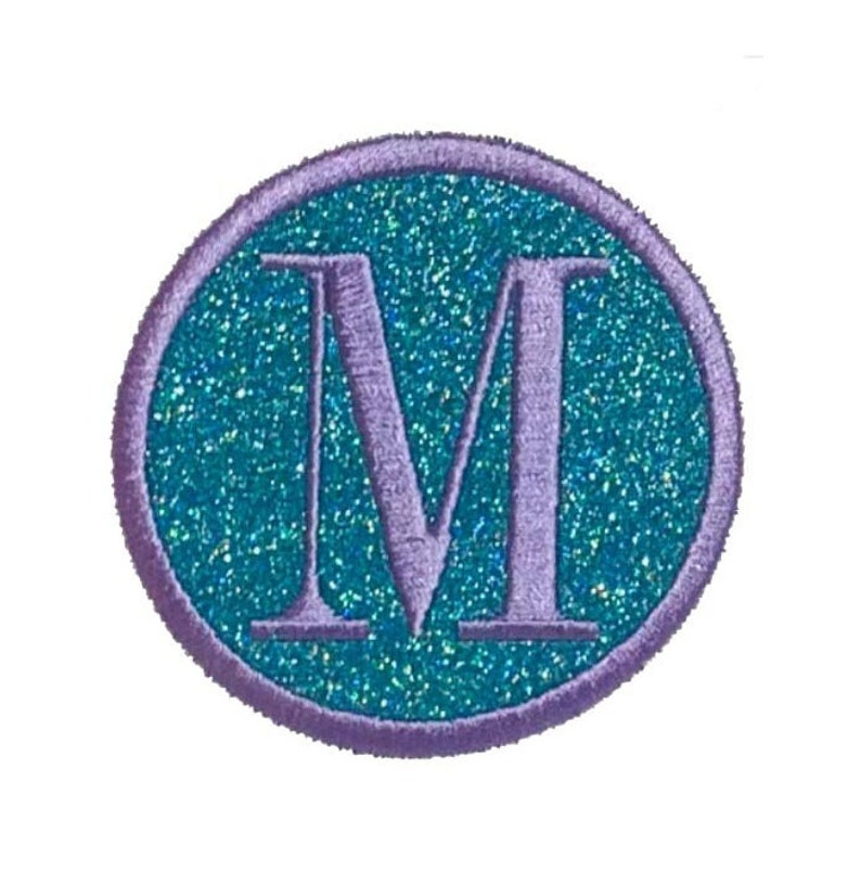 Puede incluir: Un parche de planchado de purpurina morado y turquesa con la letra "M" en blanco.