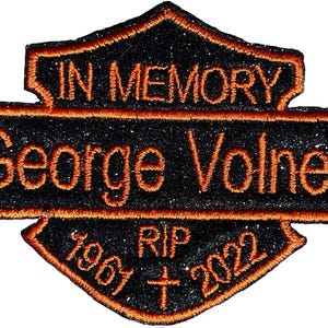 Pode incluir: Um patch bordado preto e laranja com as palavras "IN MEMORY" e "George Volner" em fio laranja. O patch também inclui os anos "1961" e "2022" com "RIP" no centro. O patch tem uma textura brilhante.