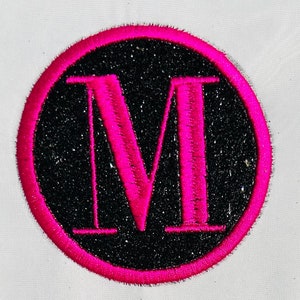 Puede incluir: Un c&iacute;rculo rosa con un fondo negro brillante y una letra M rosa en el centro.