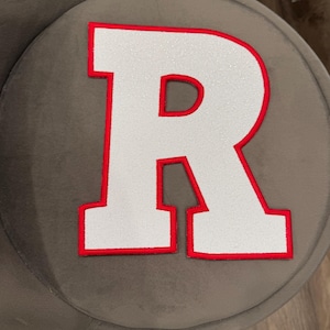 May include: White glitter-filled letter "R" with a red border. The letter is on a grey, round surface. The letter is a bold, block style.
