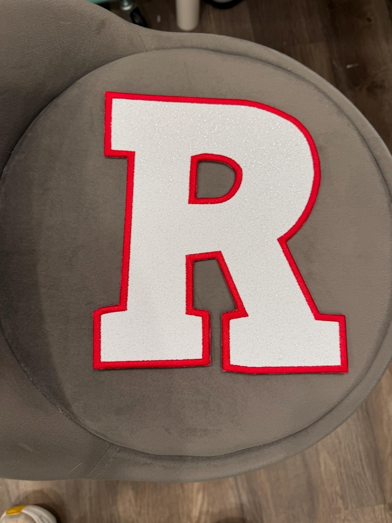 May include: White glitter-filled letter "R" with a red border. The letter is on a grey, round surface. The letter is a bold, block style.