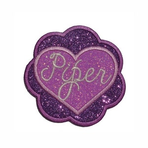 Könnte beinhalten: Ein lila glitzernder Aufnäher mit herzförmiger Mitte. Das Herz enthält den Namen "Piper" in Schreibschrift. Der Aufnäher hat einen gewellten Rand und eignet sich für Kleidung oder Accessoires.