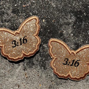 Puede incluir: Dos parches de mariposa de color dorado con purpurina con el número "3:16" impreso en ellos.