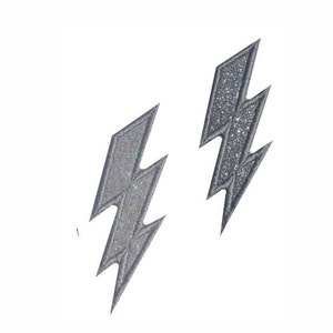 Op de afbeelding: Twee grijze, glinsterende bliksemschicht patches. Elke patch heeft een gekarteld zigzagontwerp met een getextureerd oppervlak. De patches zijn geïsoleerd op een witte achtergrond, die hun vorm en details benadrukt.