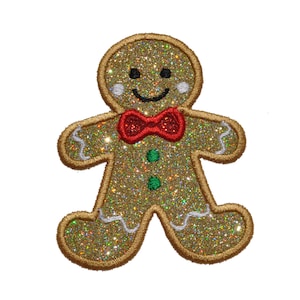 以下が含まれることがあります： 赤い蝶ネクタイと緑のボタンが付いた、金色のグリッターで覆われたジンジャーブレッドマン。ジンジャーブレッドマンは笑顔で、縁には白いアイシングが施されています。